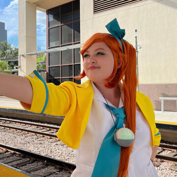 A waist-up photograph of Sailor cosplaying as Athena Cykes in her objection pose, arm outstretched and smiling triumphantly. She wears her usual yellow court outfit. The background shows train tracks, some buildings, and a cloudy blue sky.